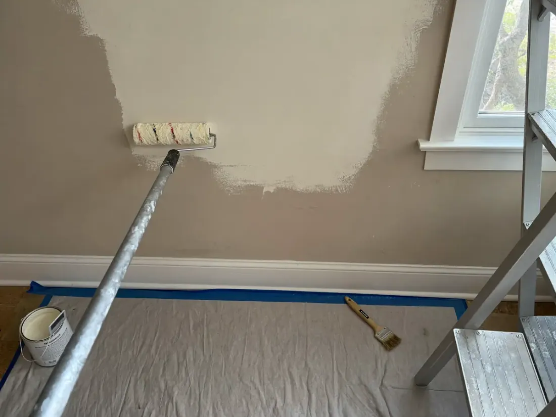 Wall being painted with a roller and painter's tape along the baseboard